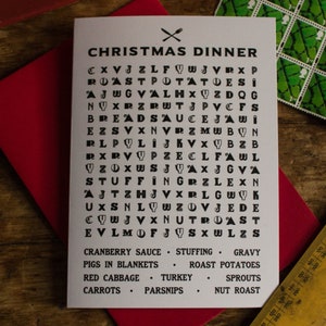 Puede incluir: Una tarjeta navide&ntilde;a con un juego de b&uacute;squeda de palabras "Cena de Navidad" sobre un sobre rojo. La tarjeta presenta una cuadr&iacute;cula de letras con palabras como "Pavo" y "Salsa" ocultas. Cubiertos cruzados en la parte superior.