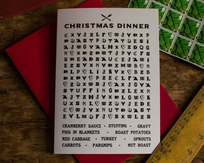 Puede incluir: Una tarjeta navide&ntilde;a con un juego de b&uacute;squeda de palabras "Cena de Navidad" sobre un sobre rojo. La tarjeta presenta una cuadr&iacute;cula de letras con palabras como "Pavo" y "Salsa" ocultas. Cubiertos cruzados en la parte superior.