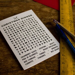 Puede incluir: Un rompecabezas de b&uacute;squeda de palabras de Cena de Navidad en blanco con texto negro, con alimentos festivos. Una regla amarilla, un bol&iacute;grafo azul y un l&aacute;piz sobre una superficie de madera.