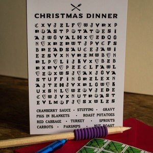 Puede incluir: Una tarjeta blanca de cena de Navidad con un juego de b&uacute;squeda de palabras y una lista de alimentos festivos. Un l&aacute;piz y un bol&iacute;grafo azul descansan sobre un sobre rojo con un sello verde.