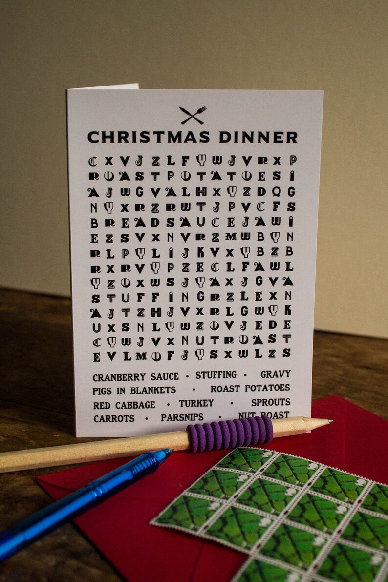 Puede incluir: Una tarjeta blanca de cena de Navidad con un juego de b&uacute;squeda de palabras y una lista de alimentos festivos. Un l&aacute;piz y un bol&iacute;grafo azul descansan sobre un sobre rojo con un sello verde.