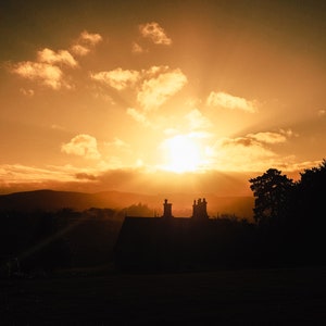 May include: A photograph of a sunset over a landscape. The sky is a warm golden colour, with clouds and the sun's rays breaking through. Silhouetted against the light are trees, a house, and hills in the distance.