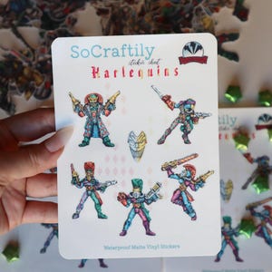 May include: A sheet of waterproof matte vinyl stickers featuring colorful Harlequins. The stickers showcase detailed illustrations of characters with weapons. The text "So Craftily" and "Harlequins" is visible.