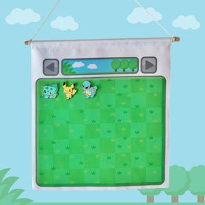 May include: A white fabric wall banner featuring a green checkered field graphic with a top interface panel containing directional arrows. Three small enamel pins of cartoon creatures are attached to the fabric, including a blue bulbous figure, a yellow mouse-like character, and a blue turtle wearing sunglasses.