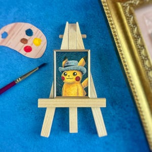 May include: A framed painting of a yellow cartoon character wearing a blue hat, displayed on a miniature wooden easel. A wooden artist's palette with paint and a paintbrush are next to the painting. The background is a textured blue surface.