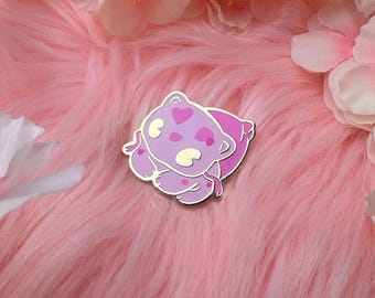 Könnte beinhalten: Ein lila Emaille-Pin mit einem Cartoon-Bären auf einem Kissen. Der Bär hat herzförmige Markierungen und einen goldenen Umriss. Der Pin befindet sich vor einem rosa, flauschigen Hintergrund mit rosa Blumen.