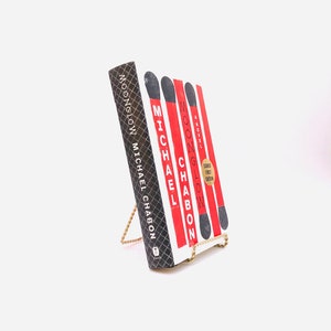May include: A red, white, and black hardcover book titled "Moonglow" by Michael Chabon. The book is on a gold metal book stand.