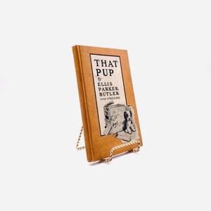 Pode incluir: Um livro castanho intitulado "That Pup" de Ellis Parker Butler, autor de "Pigs is Pigs", com uma ilustração a preto e branco de um cão na capa. O livro está num suporte de metal dourado.