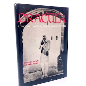 May include: A black and white photo of a man in a dark suit standing in front of a white building. The book cover is titled "In Search of Dracula: A True History of Dracula and Vampire Legends" by Raymond T. McNally and Radu Florescu.