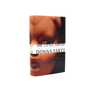 Könnte beinhalten: Ein Hardcover-Buch mit dem Titel "The Little Friend" von Donna Tartt. Das Buchcover zeigt eine Nahaufnahme eines Puppengesichts in Braun- und Schwarztönen, mit dem Titel und dem Namen des Autors in weißem Text.