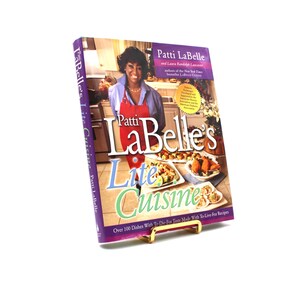 Puede incluir: Un libro de cocina titulado "Patti LaBelle's Lite Cuisine" se muestra en un atril dorado. La portada presenta a Patti LaBelle con un delantal rojo, con imágenes de comida. El título del libro está en letras grandes y coloridas.