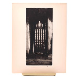 May include: A black and white print of a cathedral interior, featuring a large arched window with detailed panes. The print is mounted on a cream-colored paper, and the image is held by a gold-colored stand.