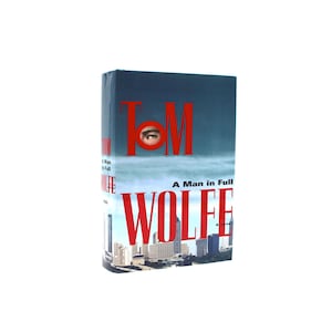 May include: A hardcover book titled "A Man in Full" by Tom Wolfe. The cover features a cityscape with the author's name in large red letters and an eye peering through the "O" in "TOM".