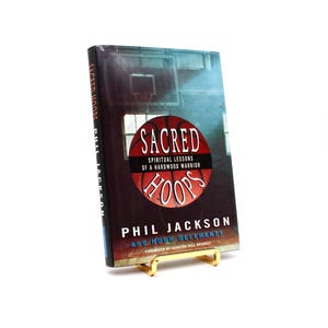 Puede incluir: Un libro de tapa dura titulado "Sacred Hoops: Spiritual Lessons of a Hardwood Warrior" de Phil Jackson y Hugh Delehanty. La portada presenta un balón de baloncesto con la palabra "Sacred" encima y "Hoops" debajo.