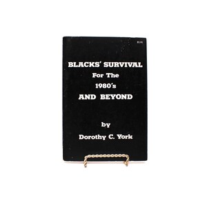 May include: A black book titled "BLACKS' SURVIVAL For The 1980's AND BEYOND" by Dorothy C. York. The book is displayed on a gold-coloured wire stand against a white background. The book's cover has white text.