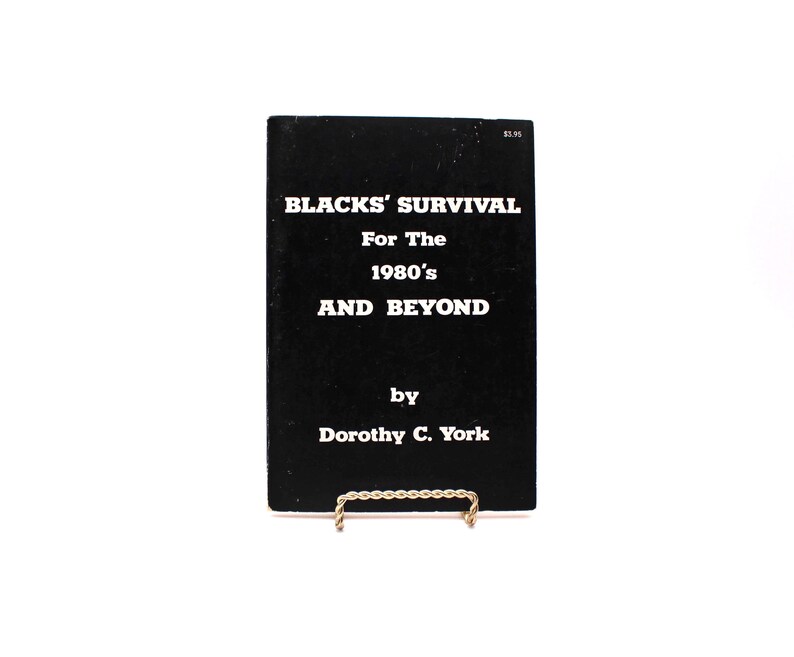 May include: A black book titled "BLACKS' SURVIVAL For The 1980's AND BEYOND" by Dorothy C. York. The book is displayed on a gold-coloured wire stand against a white background. The book's cover has white text.