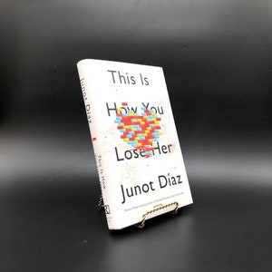 Puede incluir: Un libro blanco con el título "This Is How You Lose Her" de Junot Díaz. La portada del libro presenta un diseño de corazón pixelado en rojo, amarillo, azul y verde.