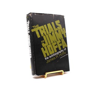 Puede incluir: Un libro negro de tapa dura titulado "The Trials of Jimmy Hoffa: The Autobiography of James R. Hoffa". El título está en letras grandes, amarillas y en negrita.