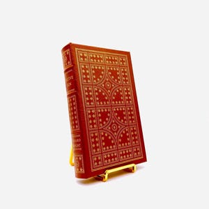 Può includere: Un libro rilegato in pelle rossa con lettere dorate e un motivo decorativo sulla copertina. Il libro è intitolato "The Narrative of Arthur Gordon Pym" ed è di Edgar Allan Poe. Il libro è su un supporto in metallo dorato.