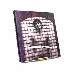 May include: A hardcover book titled "Mapplethorpe Altars" is propped up by a gold-colored book stand. The book cover features a black and white image of a person behind a metal grid, with a purple border.