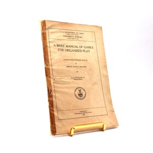 Könnte beinhalten: Ein Vintage-Handbuch mit dem Titel "A Brief Manual of Games for Organized Play" von Martha Travella Speakman. Das Cover ist in verblichenem Beige mit schwarzem Text und einem Siegel gehalten. Das Buch steht auf einem goldfarbenen Ständer.