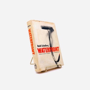Puede incluir: Un libro de bolsillo vintage titulado "Waterfront" de Budd Schulberg. La portada presenta una ilustración en blanco y negro de un gancho.