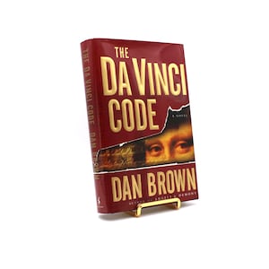 May include: A red hardcover book with gold lettering titled "The Da Vinci Code" by Dan Brown. The book cover features a close-up of a woman's eye.