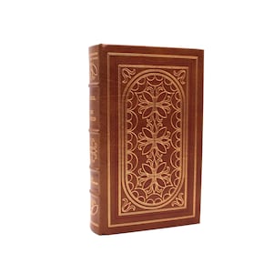 May include: A brown hardcover book with gold foil stamping on the front cover and spine. The front cover features an oval frame containing three floral motifs. The spine has raised bands with gold decorative accents and text in gold lettering.