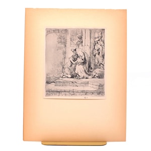 Puede incluir: Un grabado en blanco y negro enmarcado que representa una escena bíblica. La obra de arte muestra a un hombre arrodillado ante otro, con figuras en el fondo. La impresión está sobre un papel color crema, montado sobre un fondo más grande, de color naranja claro.