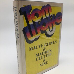 May include: A hardcover book by Tom Wolfe, titled "Mauve Gloves & Madmen, Clutter & Vine." The title "Tom Wolfe" is in a retro, multi-coloured font, set against a yellow background. The book's spine is visible, with the author's name.