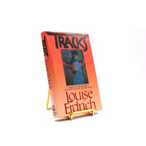 Könnte beinhalten: Ein Buch mit dem Titel "Tracks" von Louise Erdrich, präsentiert auf einem kleinen, goldfarbenen Metallständer. Das Buchcover ist orange mit dem Titel in großen schwarzen Buchstaben und einer farbenfrohen Illustration.