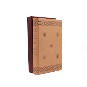 Może przedstawiać: Dwie książki w twardej oprawie. Książka po lewej stronie ma ciemnobordową okładkę. Książka po prawej stronie ma beżową okładkę z geometrycznym wzorem u góry i u dołu oraz wzorami rozbłysków gwiazd. Tekst na okładce brzmi "The Voyage of H.M.S. Beagle" i "The Journal of Charles Darwin."