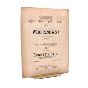 Puede incluir: Una partitura antigua titulada "Who Knows?" con texto en tinta negra sobre papel envejecido. La canción es de Ernest R. Ball, con letra de Paul Laurence Dunbar. La partitura está sostenida por un soporte dorado.