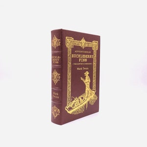 Pode incluir: Um livro encadernado em couro castanho com letras douradas na lombada e na capa. O título é "Adventures of Huckleberry Finn (Tom Sawyer's Companion)" de Mark Twain. A capa apresenta uma ilustração dourada de um homem num barco.