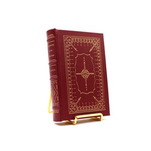 Puede incluir: Un libro de tapa dura de color granate con patrones decorativos y texto en relieve dorado. El libro se exhibe en un pequeño soporte de metal dorado. El lomo del libro presenta el título y el nombre del autor en letras doradas.