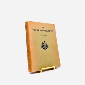 May include: A vintage paperback book titled "The Virgin and the Gipsy" by D.H. Lawrence. The book is displayed on a gold metal book stand.