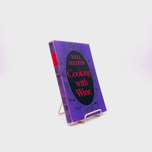 May include: A purple book titled "Cooking with Wine" by Nell Heaton. The book is on a gold metal book stand.