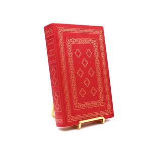 Pode incluir: Um livro encadernado em vermelho com uma borda decorativa dourada e o título "O Vermelho e o Negro" na lombada. O livro está em um suporte de livro de metal dourado.