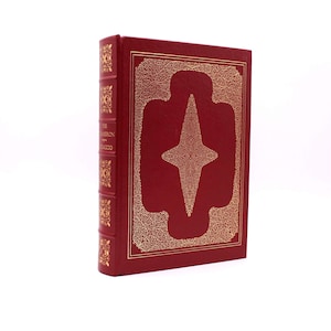 Könnte beinhalten: Ein rotes, ledergebundenes Buch mit goldenen Lettern auf dem Rücken. Der Titel lautet "The American Tragedy" von Theodore Dreiser. Der Einband zeigt ein goldenes Sternmuster.