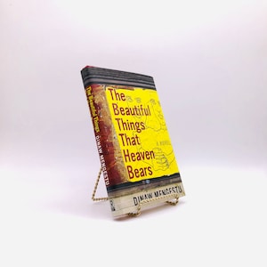 May include: A yellow paperback book with a black and white illustration of two faces and hands on the cover. The title is "The Beautiful Things That Heaven Bears" by Dinnaw Mengestu.