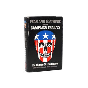 May include: A black hardcover book titled "FEAR AND LOATHING: ON THE CAMPAIGN TRAIL '72". The cover displays a skull design in red, white, and blue. The author's name and illustrator's name are printed at the bottom of the book.