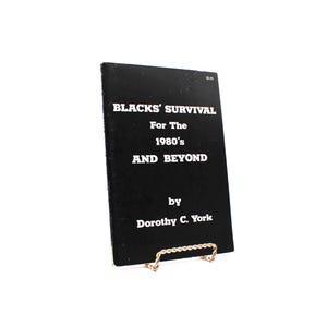 May include: A black book titled "BLACKS' SURVIVAL For The 1980's AND BEYOND" by Dorothy C. York. The book is propped up on a gold-coloured metal stand. The text is in white, and the book has a price tag in the upper right corner.