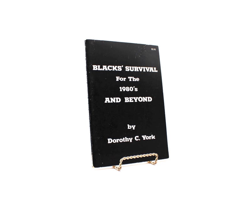 May include: A black book titled "BLACKS' SURVIVAL For The 1980's AND BEYOND" by Dorothy C. York. The book is propped up on a gold-coloured metal stand. The text is in white, and the book has a price tag in the upper right corner.