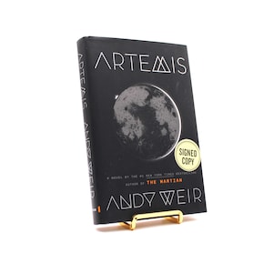 May include: A black hardcover book titled Artemis by Andy Weir, featuring a graphic of the moon on the front cover. A yellow circular sticker on the cover reads Signed Copy. The book is displayed on a small gold-colored metal stand.