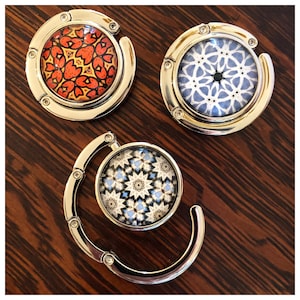 May include: Three silver-toned, foldable, circular purse hooks with different patterned designs. The first hook has a red and orange geometric pattern. The second hook has a blue and white geometric pattern. The third hook has a blue, white, and black geometric pattern.
