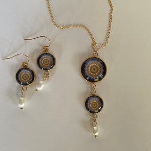 Op de afbeelding: Een goudkleurige sieradenset met een ketting en oorbellen. De ketting heeft twee ronde hangers en een bungelende parel, terwijl de oorbellen een enkele ronde hanger en een parel hebben. De hangers hebben een blauw, bruin en wit mandala-ontwerp.