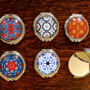 May include: Six round, silver-toned compact mirrors with different patterned designs. The mirrors are closed and feature a variety of geometric patterns in blue, red, white, and black.