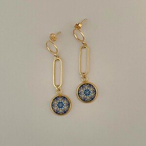 Op de afbeelding: Een paar goudkleurige oorbellen met een kleine ronde knop, een langwerpige ovale schakel en een ronde hanger. De hanger toont een gedetailleerd blauw, geel en wit geometrisch patroon in een gouden omlijsting.
