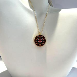 Puede incluir: Un collar de cadena dorada con un colgante redondo. El colgante presenta un fondo negro con un diseño floral en tonos rosas y naranjas. El collar se exhibe en un maniquí blanco.