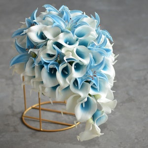 May include: A cascading bridal bouquet made of white and light blue calla lilies. The bouquet is arranged in a circular shape and is held together by a gold metal stand.